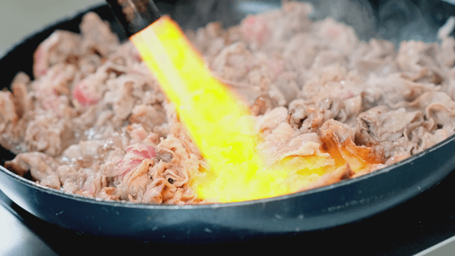 Meat being seared with a torch in a pan