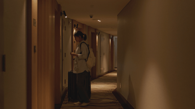 Woman walking down a hotel hallway with her suitcase into her room.