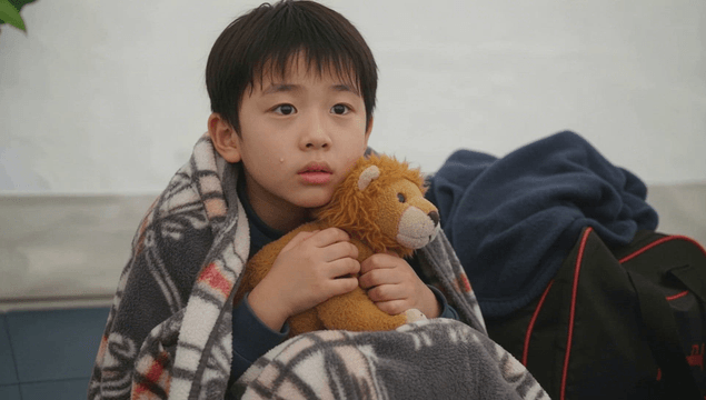 Boy wrapped in a blanket holding a lion doll in a shelter