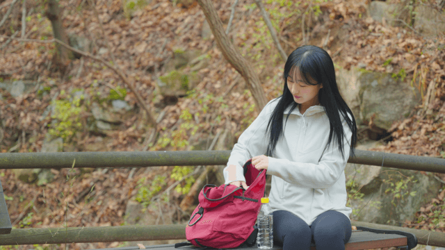 Woman placing water bottle into backpack on bench in autumn forest