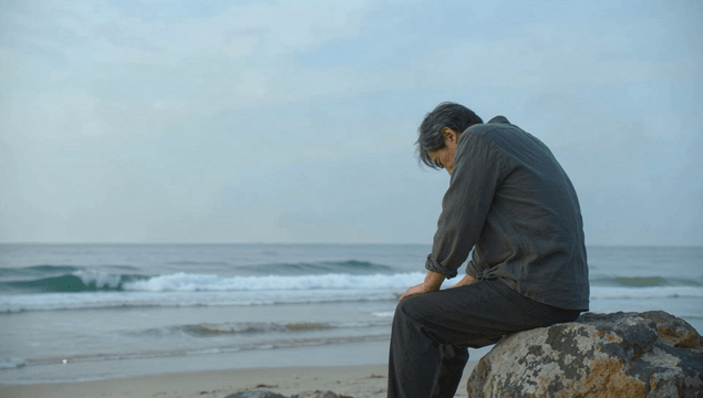 Elderly man lowering his head while sitting on rocks by the seaside