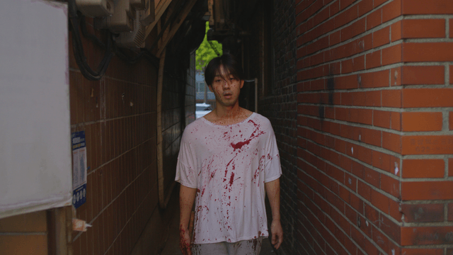 Man holding a hammer in blood-stained clothes walking down a narrow alley