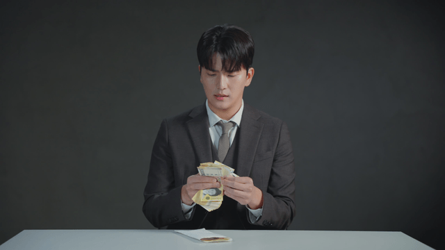 Man in a suit counting money at a desk with a dissatisfied expression