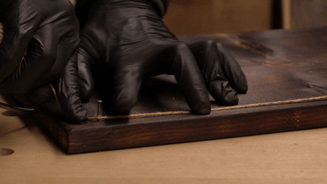 Gloved hand winding thread around a nail on wooden board