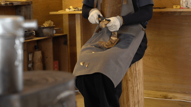 Artisan carving wood with a chisel while sitting on a chair in workshop