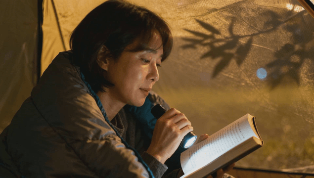 Middle-aged woman reading book with flashlight inside tent